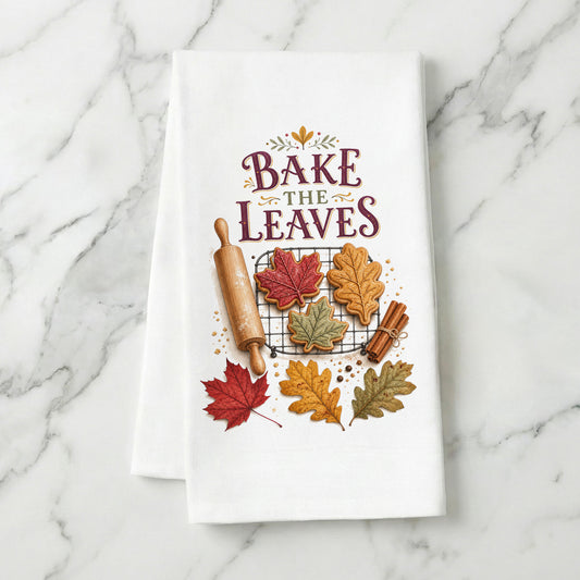 Bake the Leaves Fall Baking Tea Towel folded on white marble, with leaf-shaped cookies, cooling rack, rolling pin, cinnamon, and autumn leaves clearly shown.