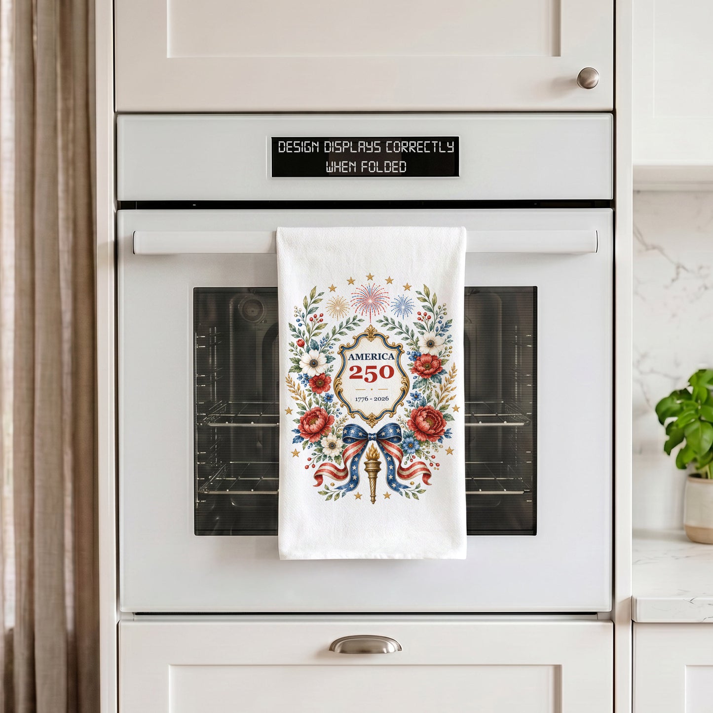 America 250 botanical wreath tea towel hanging on an oven handle, showing red white and blue floral anniversary artwork on white cotton
