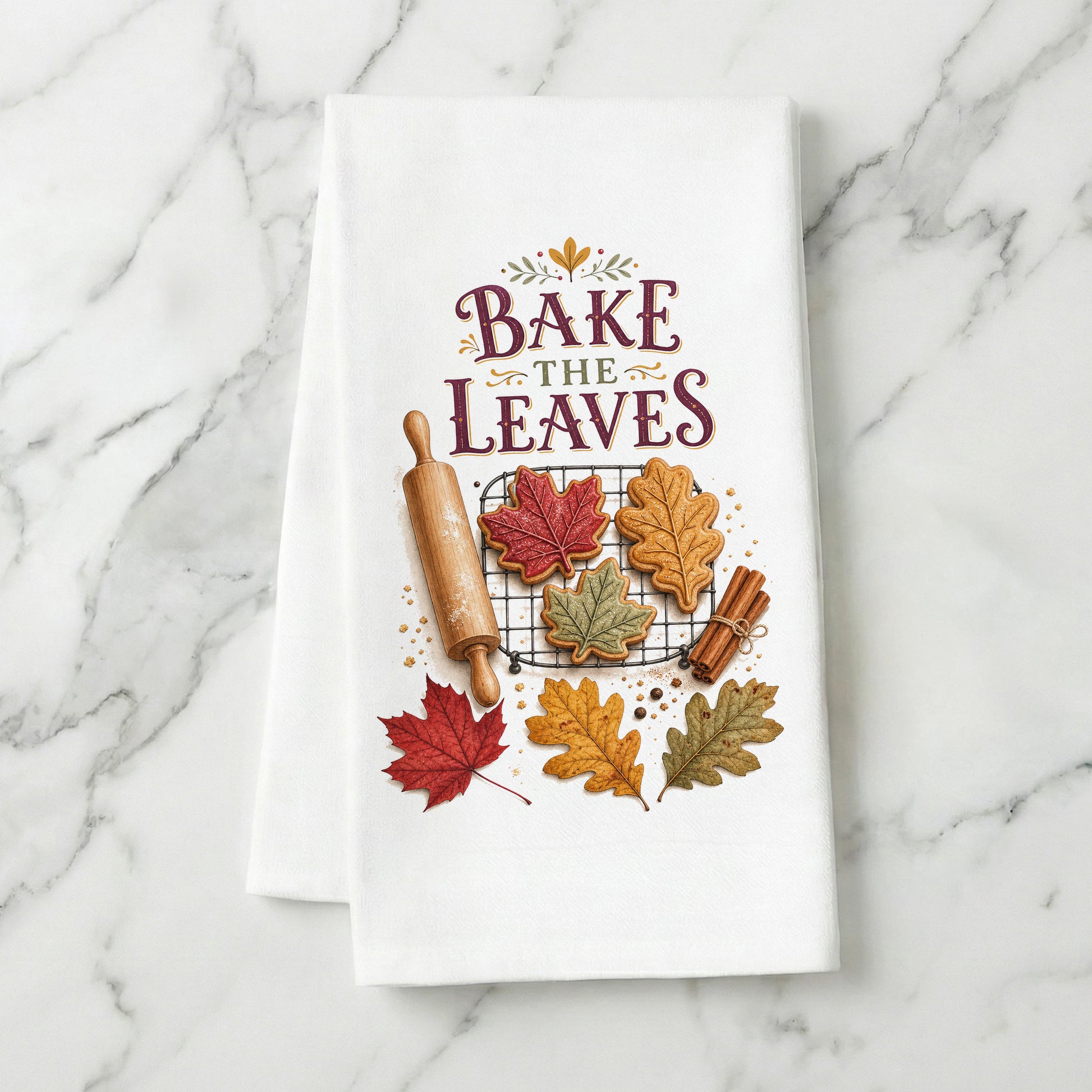 Bake the Leaves Fall Baking Tea Towel folded on white marble, with leaf-shaped cookies, cooling rack, rolling pin, cinnamon, and autumn leaves clearly shown.