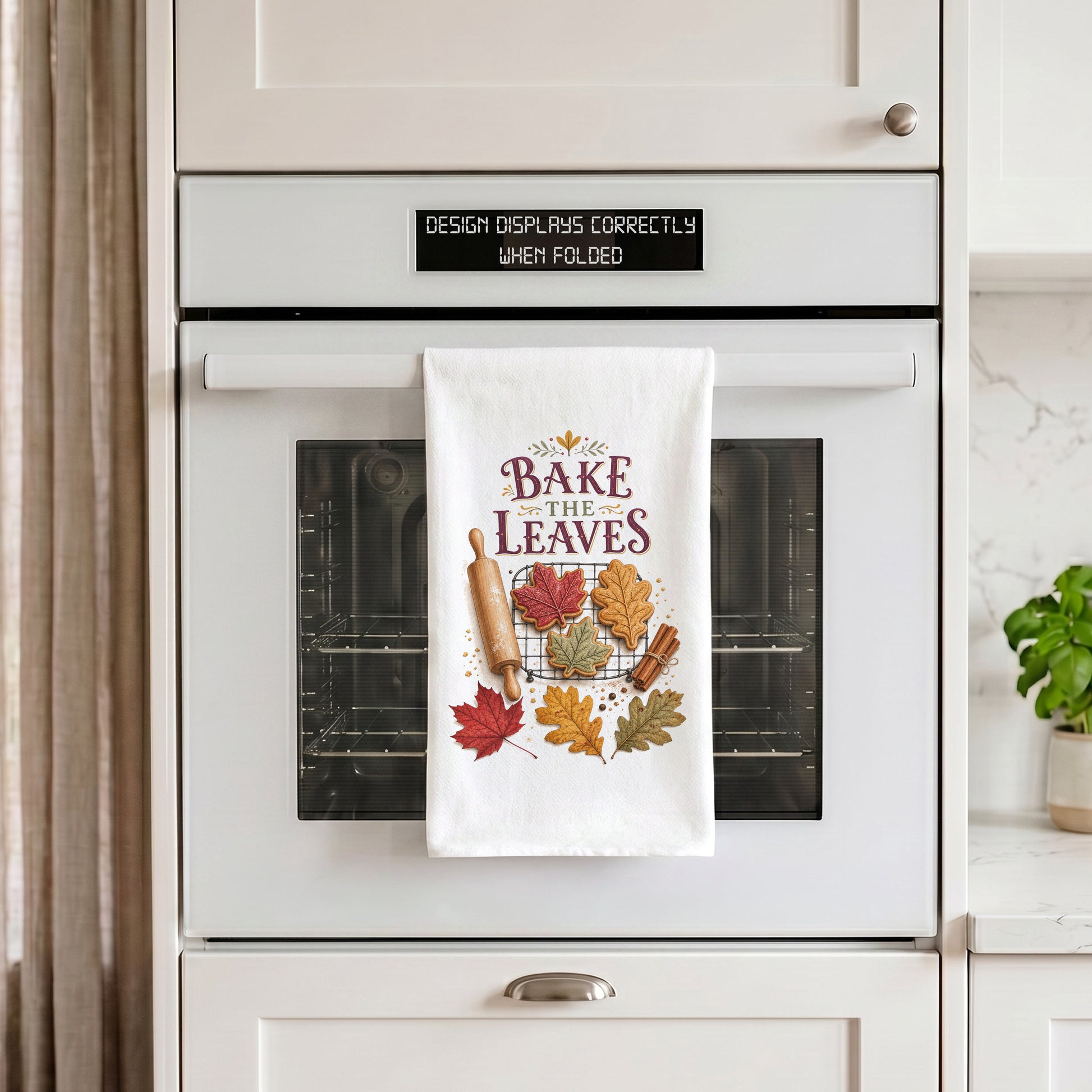 Bake the Leaves Fall Baking Tea Towel draped over an oven handle, with the artwork centered when folded.