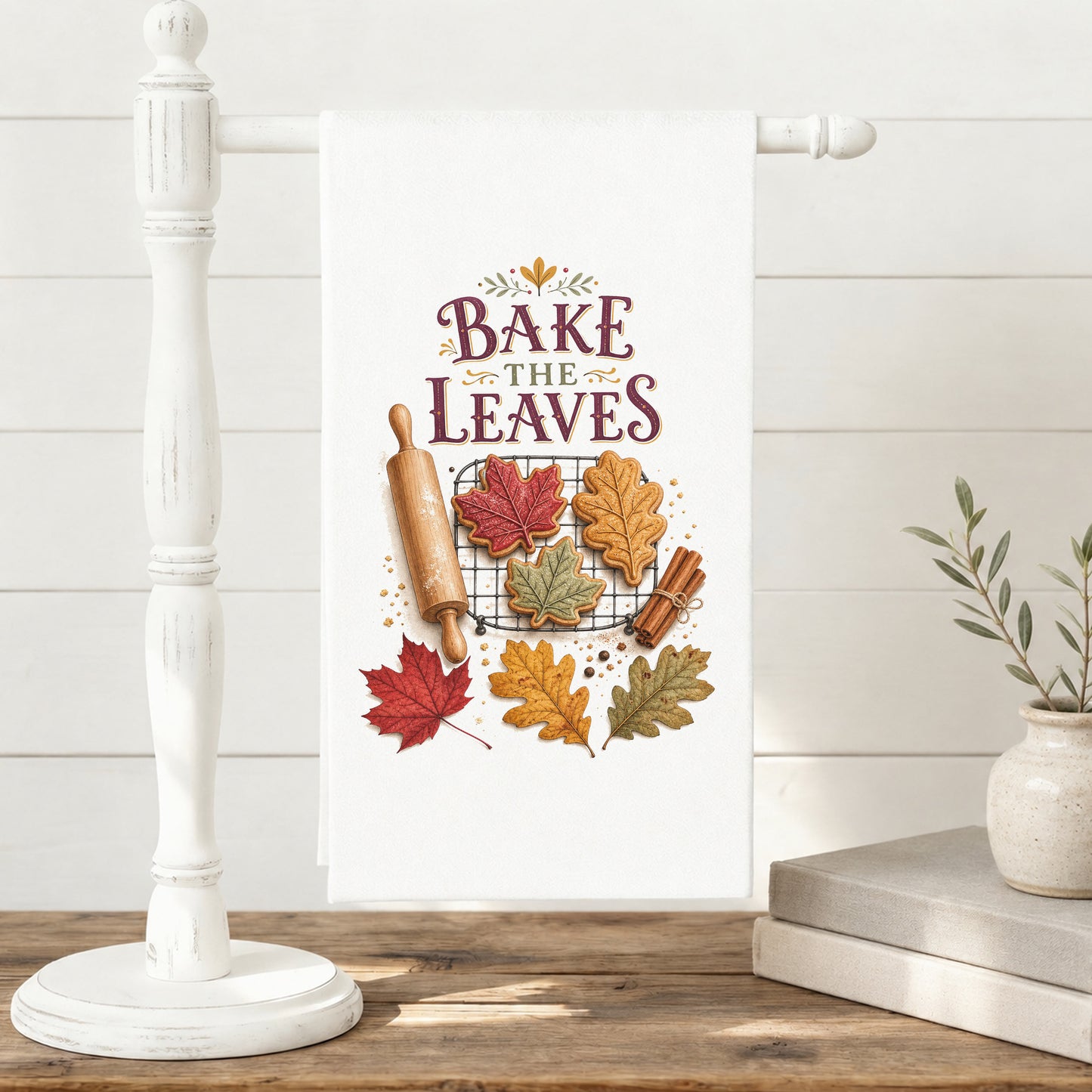 Bake the Leaves Fall Baking Tea Towel displayed on a freestanding towel stand, with leaf-shaped cookies, cooling rack, rolling pin, cinnamon, and autumn leaves clearly shown.