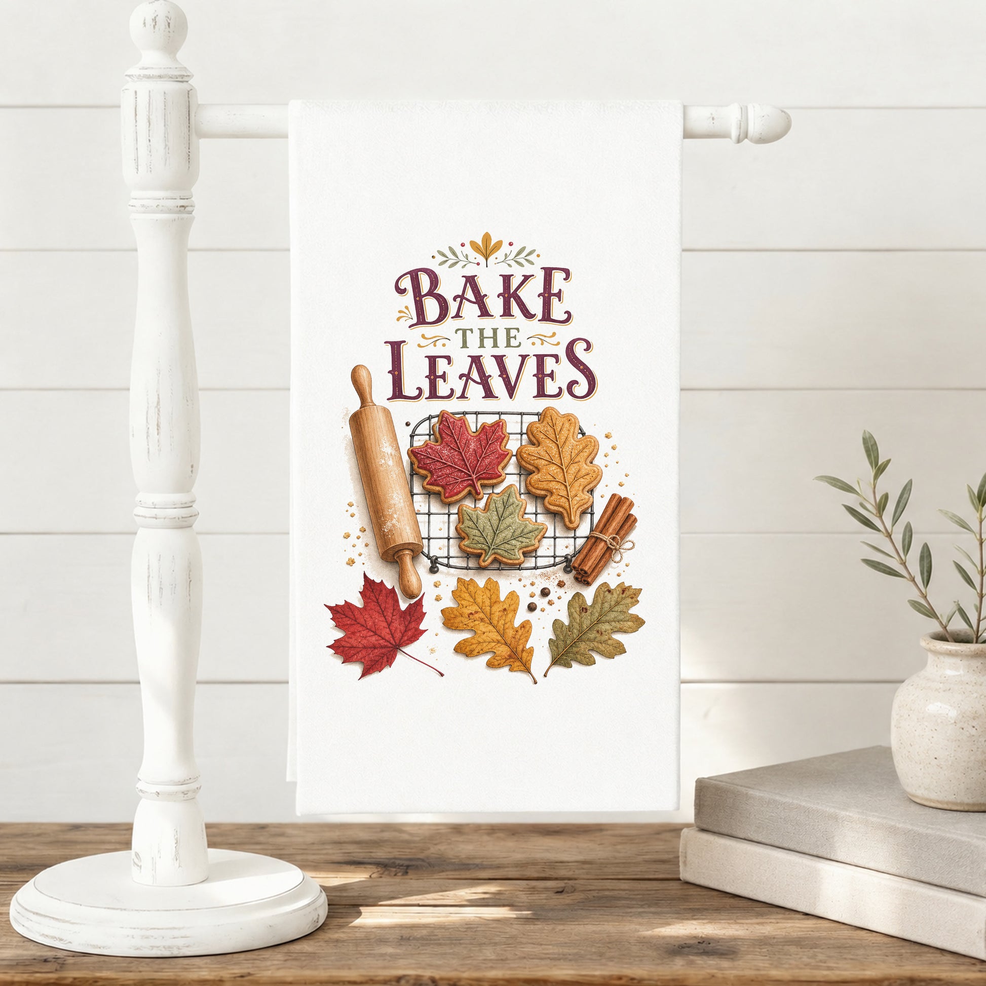 Bake the Leaves Fall Baking Tea Towel displayed on a freestanding towel stand, with leaf-shaped cookies, cooling rack, rolling pin, cinnamon, and autumn leaves clearly shown.