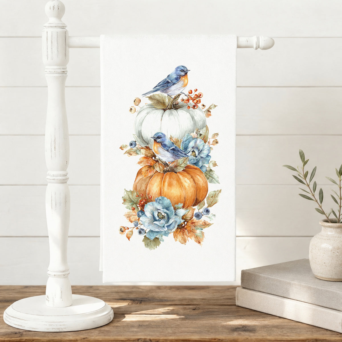 Bluebird Pumpkin tea towel displayed on a white freestanding towel stand beside books and a vase.