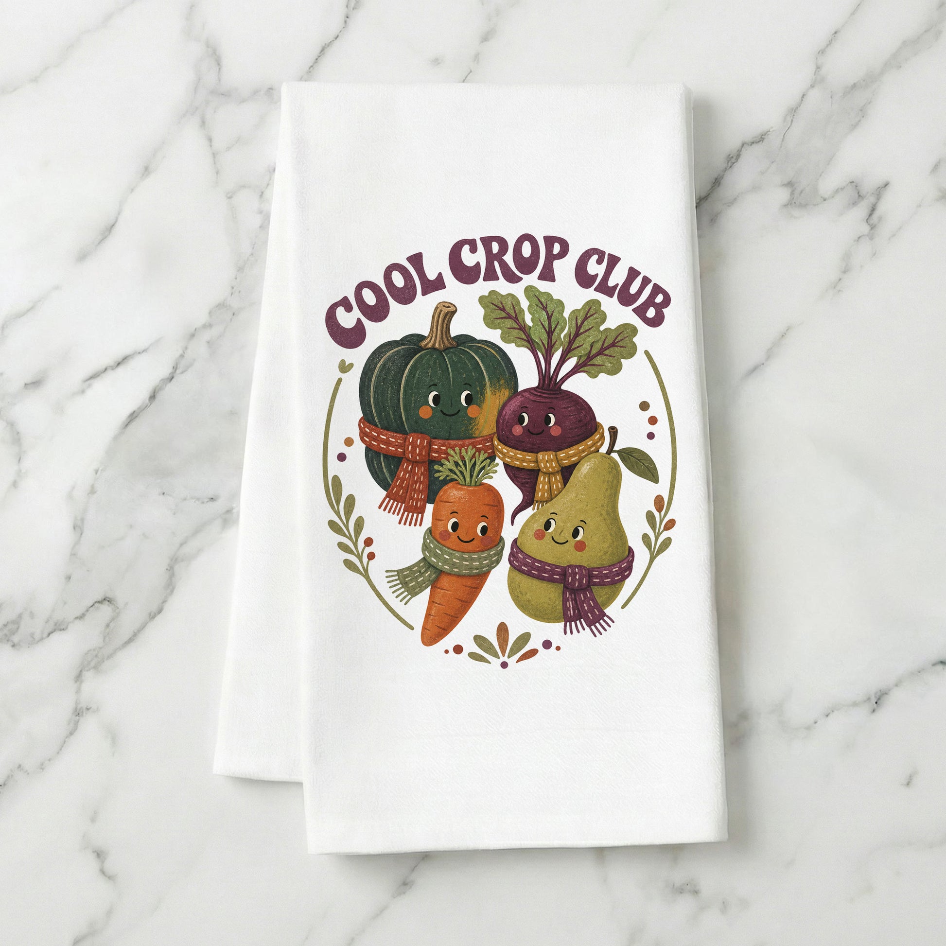 Cool Crop Club Harvest Tea Towel folded on white marble, with smiling squash, beet, carrot, and pear characters wearing scarves clearly shown.