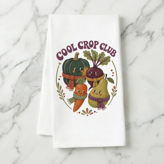 Cool Crop Club Harvest Tea Towel folded on white marble, with smiling squash, beet, carrot, and pear characters wearing scarves clearly shown.