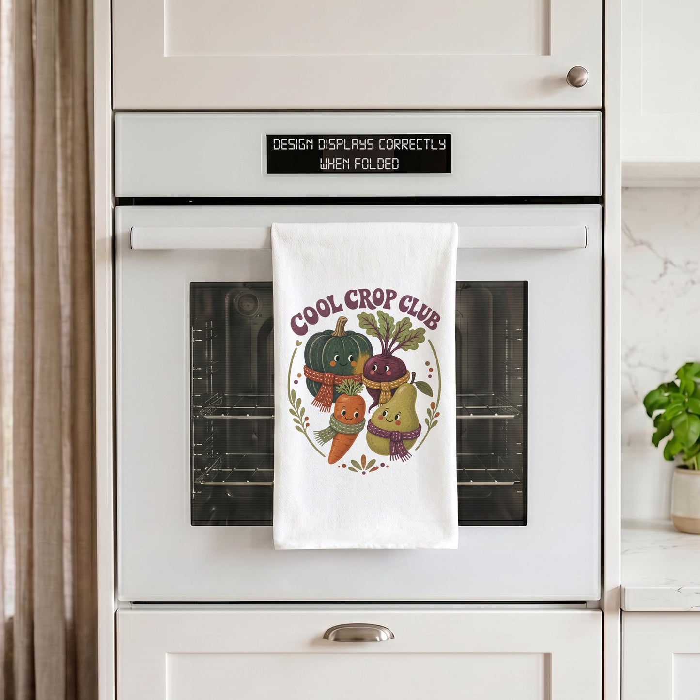 Cool Crop Club Harvest Tea Towel draped over an oven handle, with the artwork centered when folded.