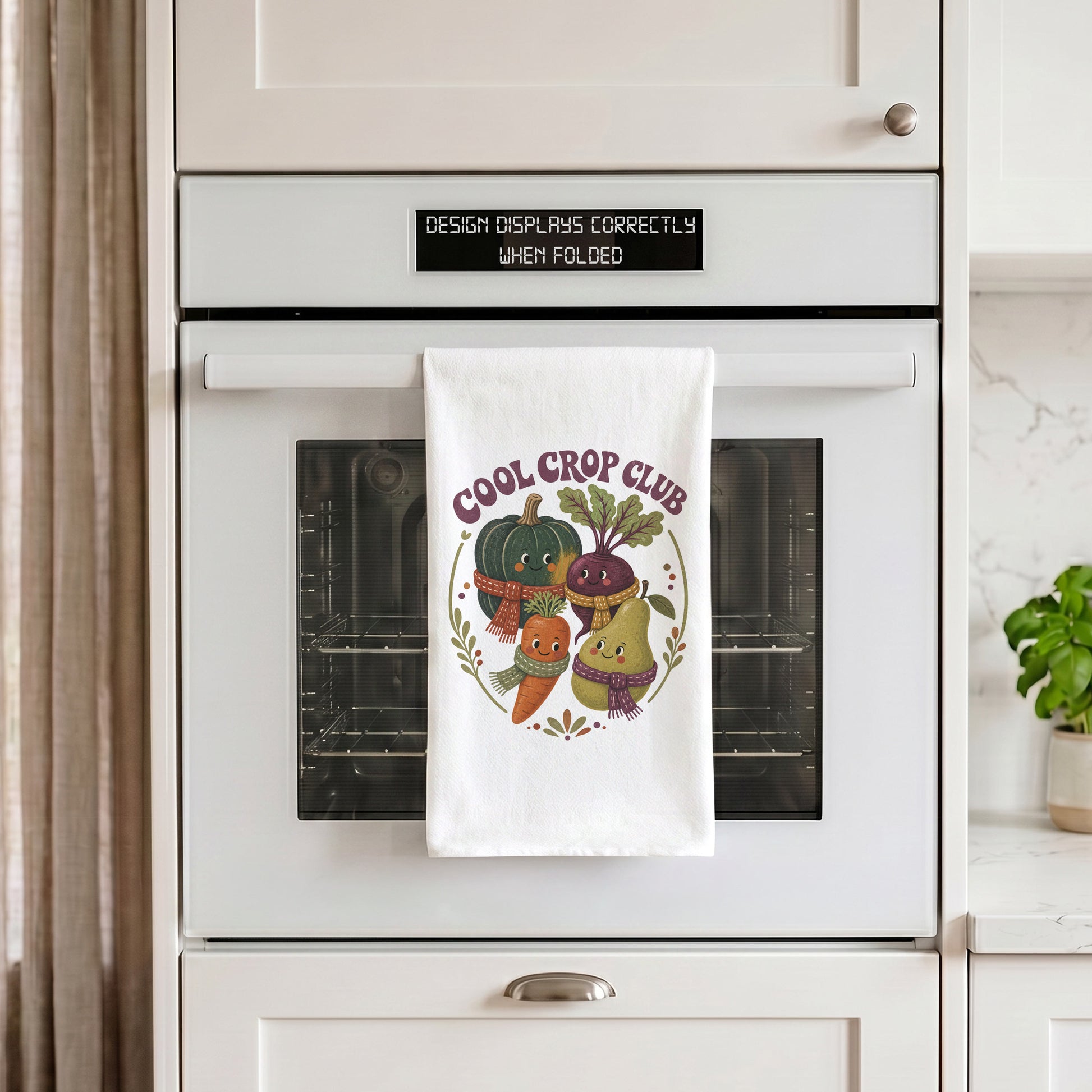 Cool Crop Club Harvest Tea Towel draped over an oven handle, with the artwork centered when folded.