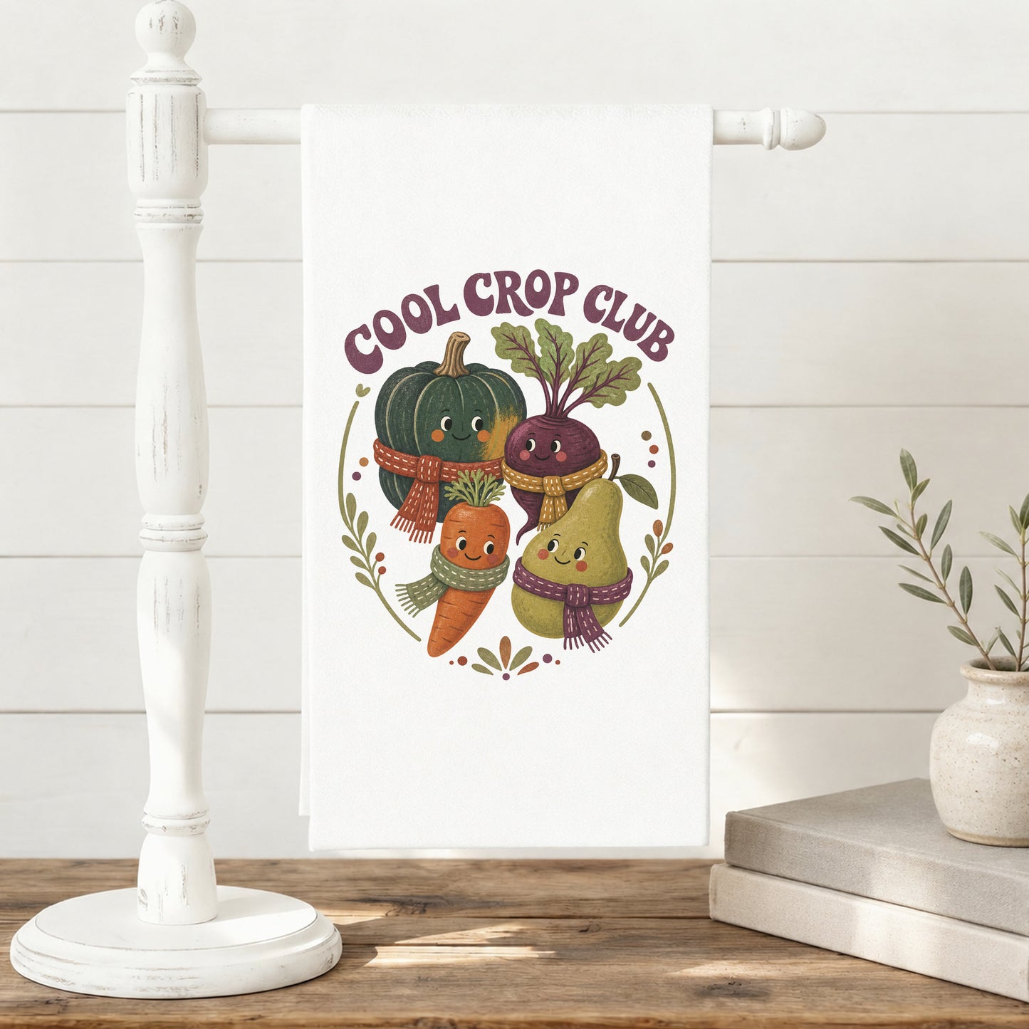 Cool Crop Club Harvest Tea Towel displayed on a freestanding towel stand, with smiling squash, beet, carrot, and pear characters wearing scarves clearly shown.
