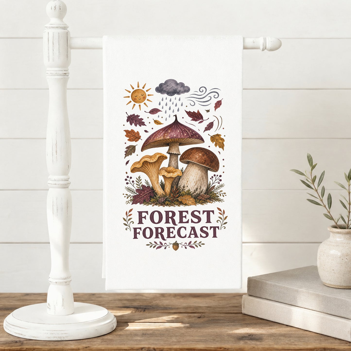 Forest Forecast Mushroom Tea Towel displayed on a freestanding towel stand, with woodland mushrooms, autumn leaves, sun, rain cloud, wind, and burgundy lettering clearly shown.