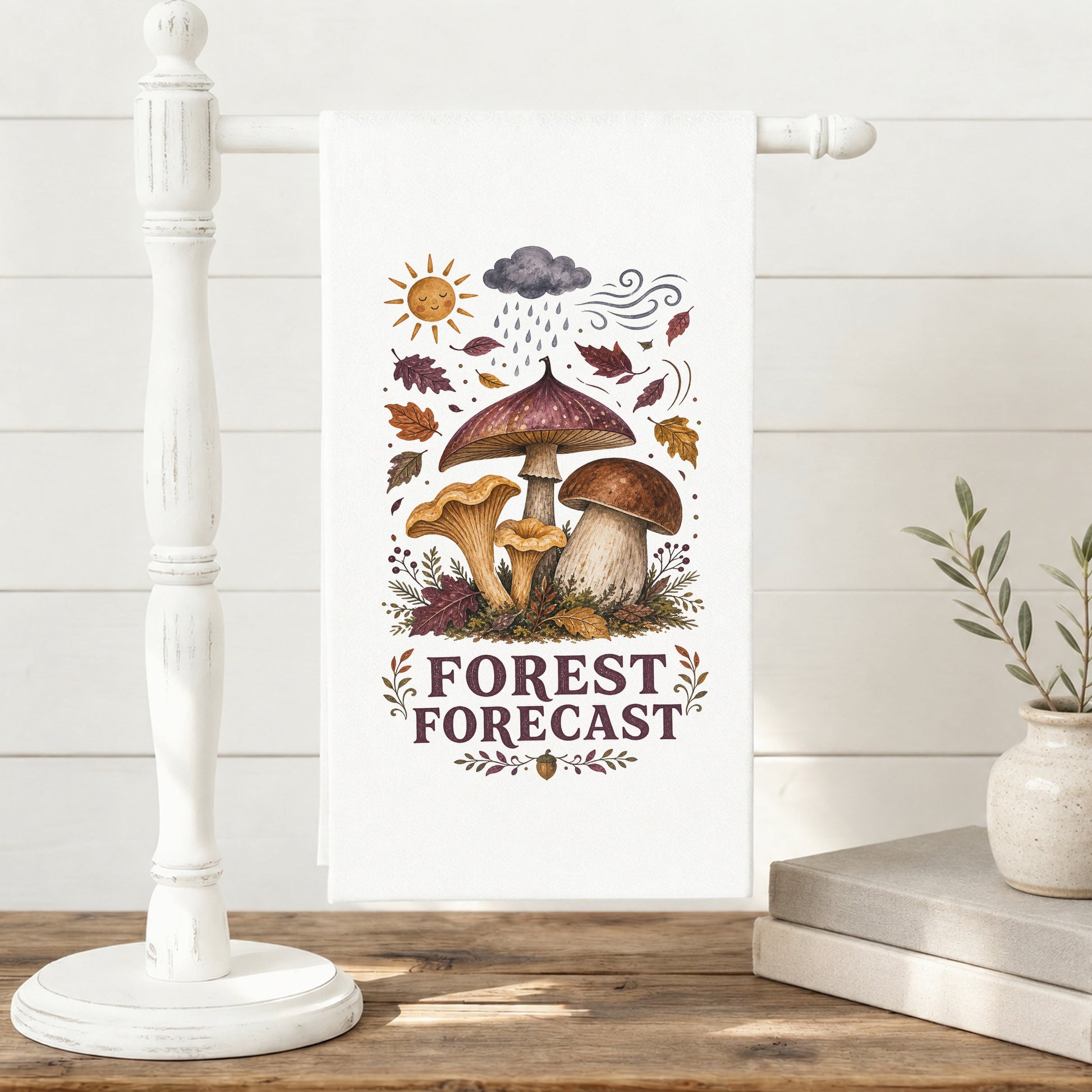 Forest Forecast Mushroom Tea Towel displayed on a freestanding towel stand, with woodland mushrooms, autumn leaves, sun, rain cloud, wind, and burgundy lettering clearly shown.