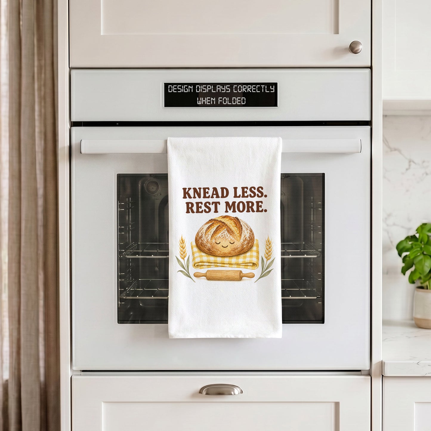 White Knead Less Rest More tea towel draped over an oven handle in a kitchen setting.