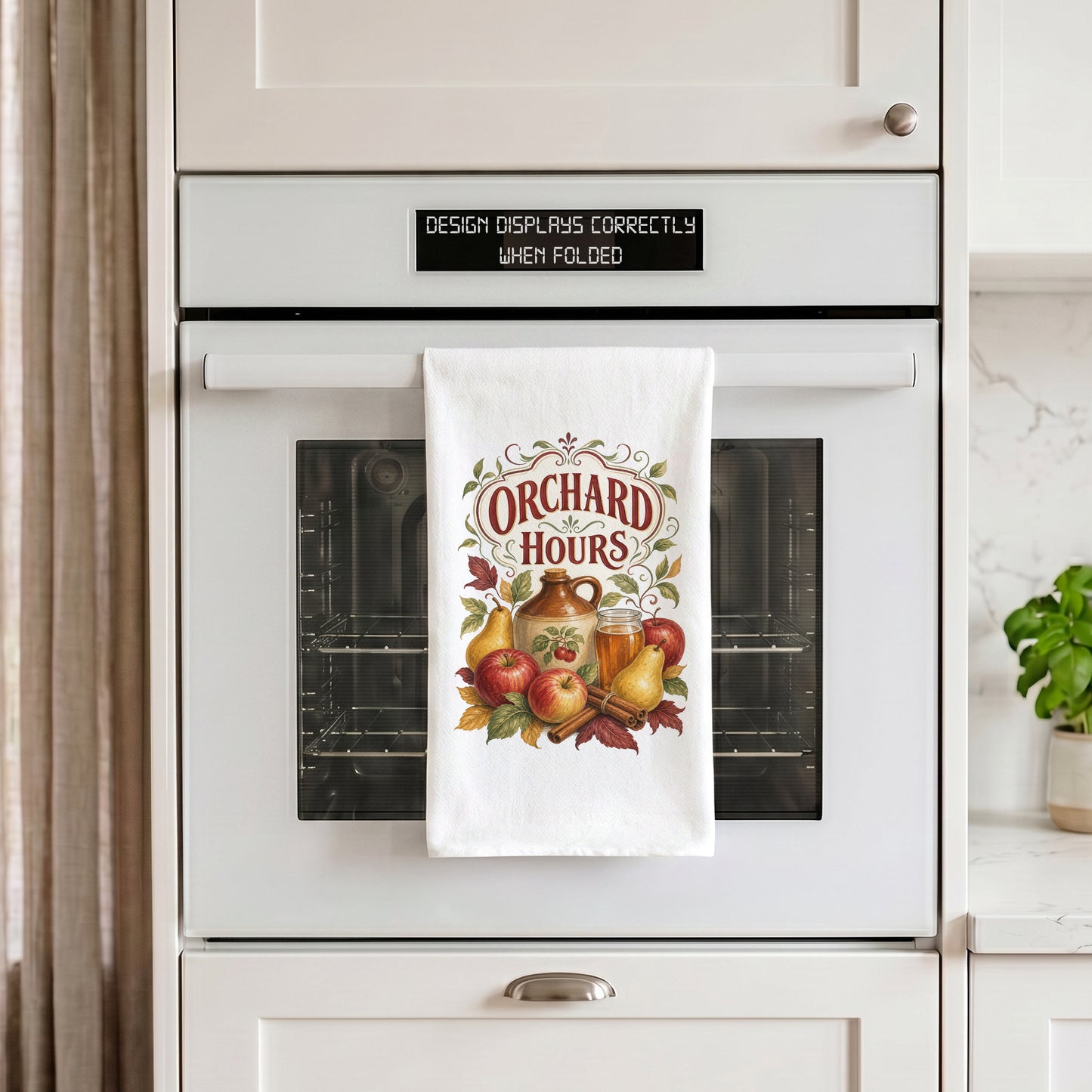 Orchard Hours Apple Cider Tea Towel draped over an oven handle, with the artwork centered when folded.