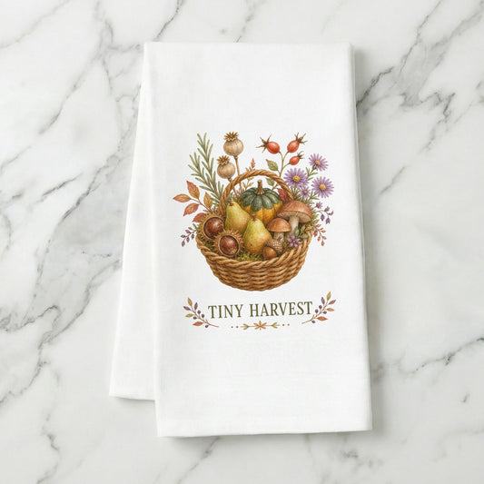 Tiny Harvest Autumn Tea Towel folded on white marble, with woven basket filled with pears, squash, mushrooms, chestnuts, herbs, and flowers clearly shown.